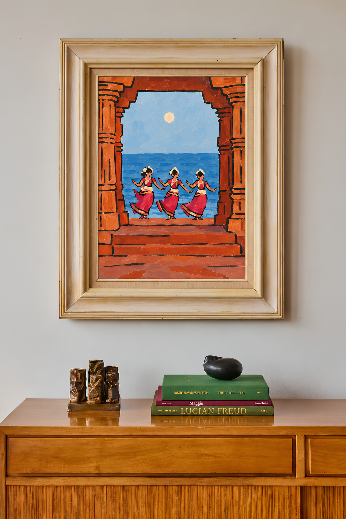 Framed stone-arch seascape painting on a wall above a wooden sideboard with books and small sculptures