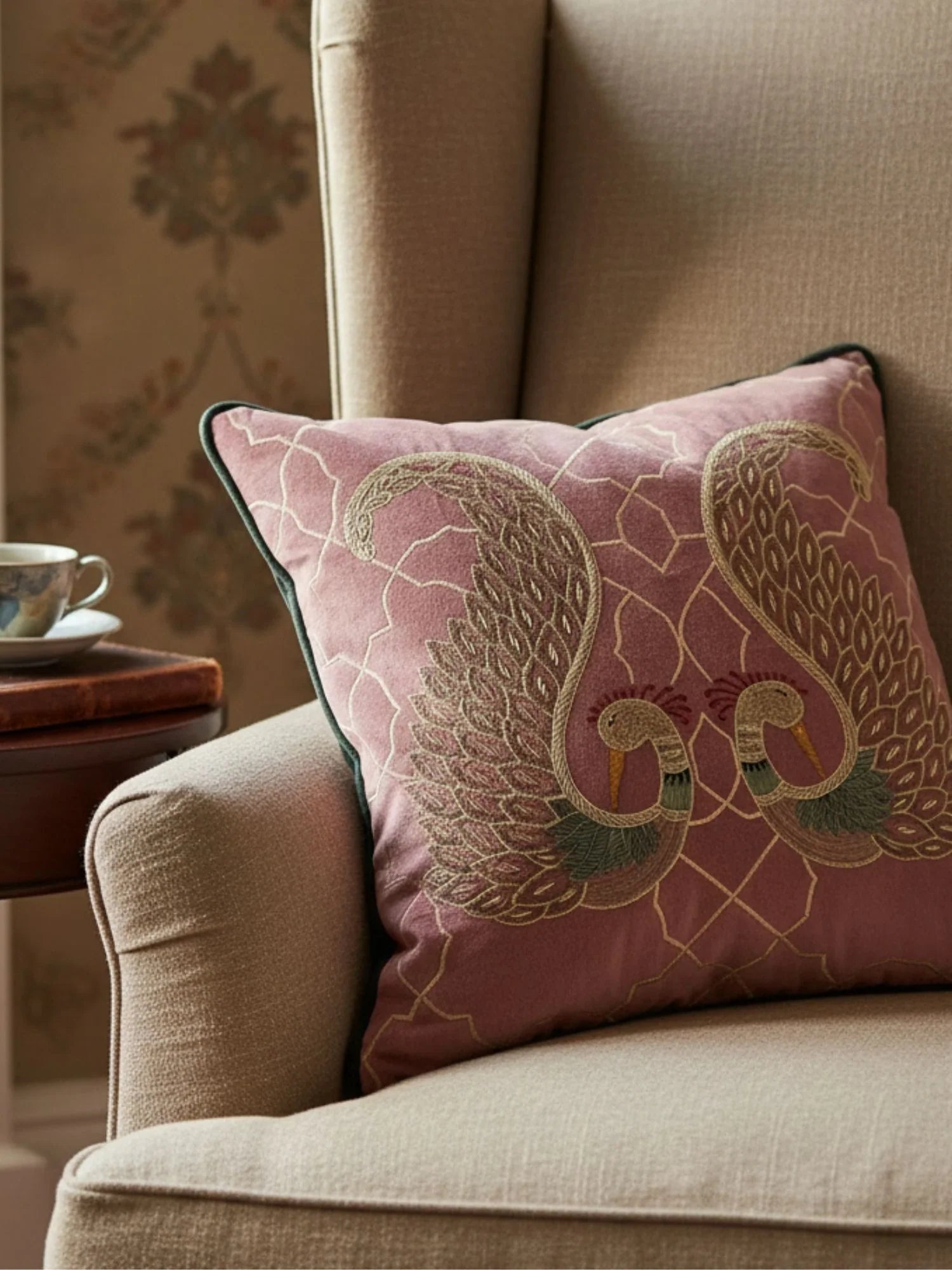 The Kashmir cushion featuring hand-embroidered bird motifs styled on a classic beige wingback chair with a side table.