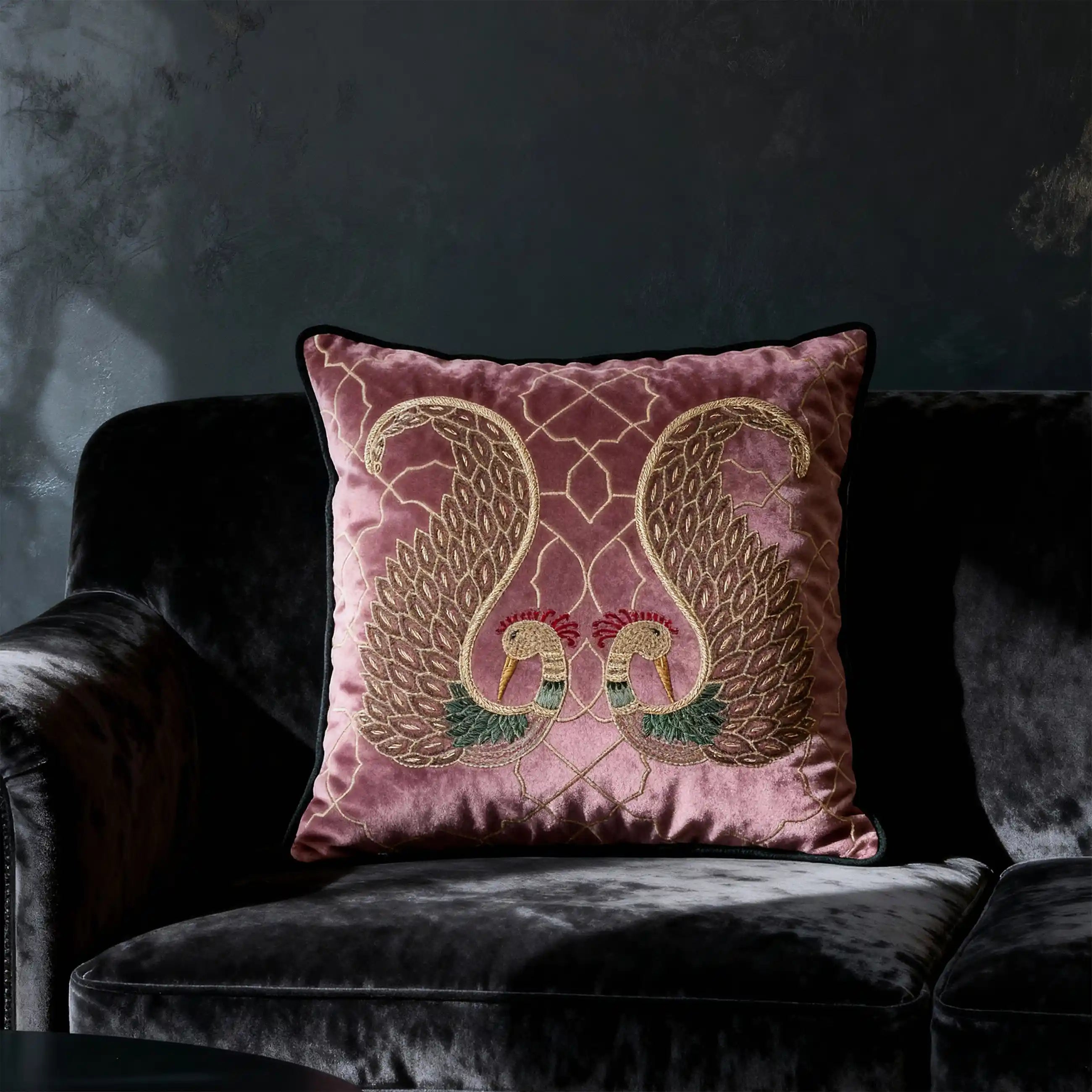 A moody interior shot of the Mauve Rose peacock cushion on a dark charcoal velvet sofa with dramatic side lighting.