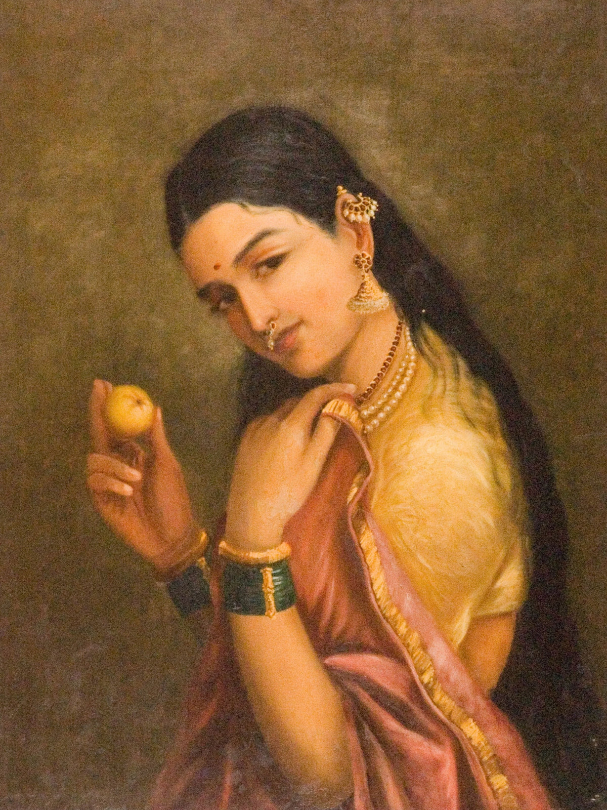 Portrait of a woman with long dark hair and gold jewellery holding a yellow fruit, in a pink sari