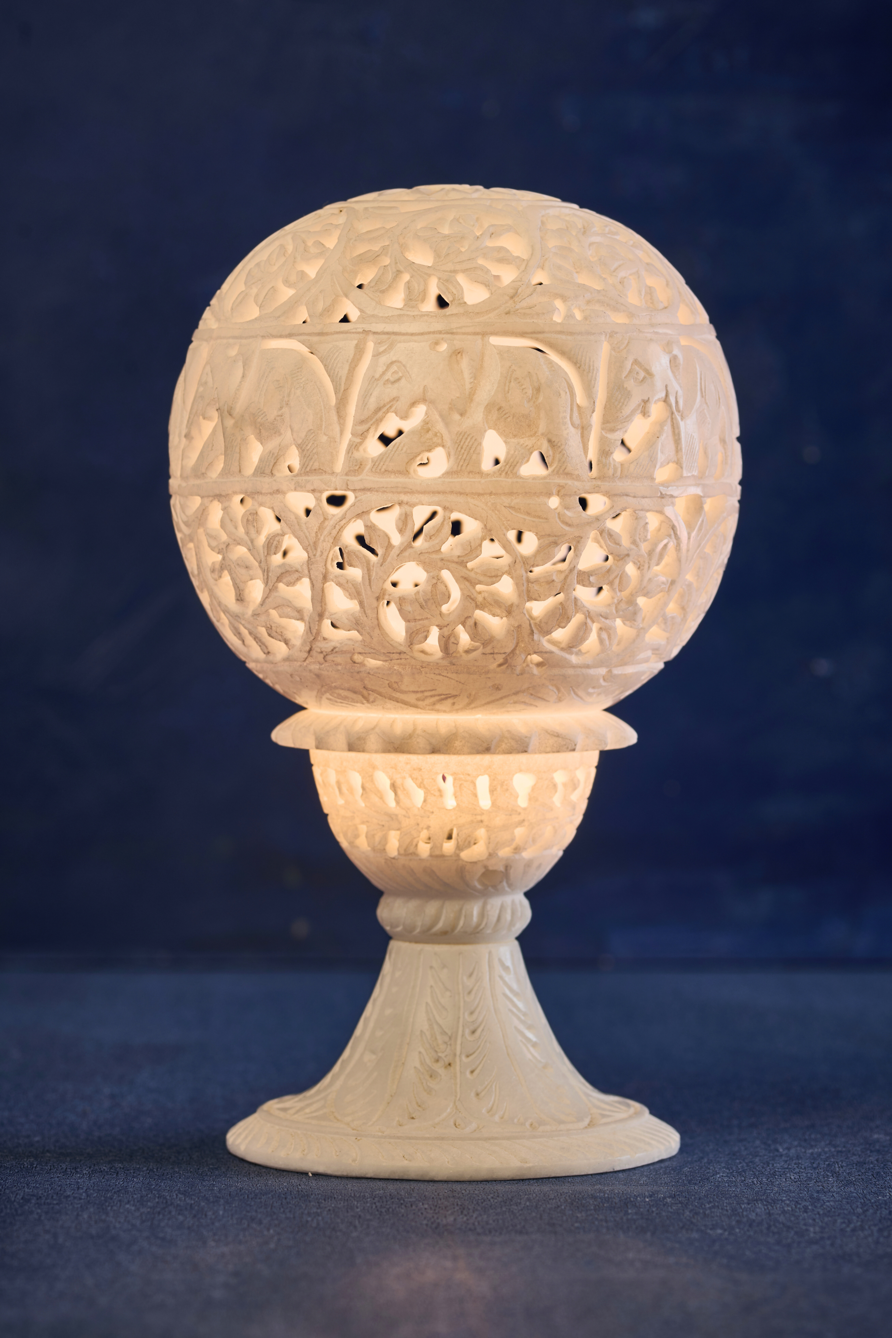 A white soapstone lampshade with intricate carvings, illuminated from within, showcasing patterns of leaves and vines.